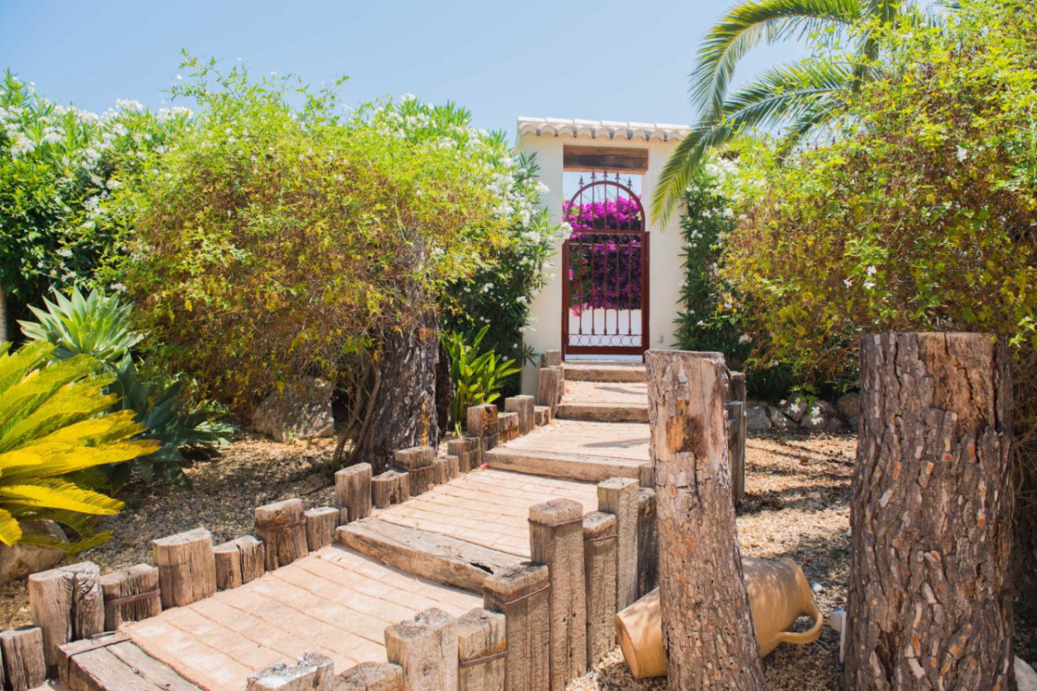 Sendero de madera y piedra rodeado de vegetación mediterránea que conduce a un portal blanco con puerta de forja y buganvillas en flor.
