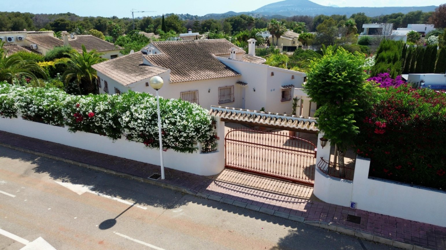 Vista exterior de una villa mediterránea con fachada blanca, tejado de terracota y puerta de hierro, rodeada de setos de adelfas en flor y vistas a la montaña.