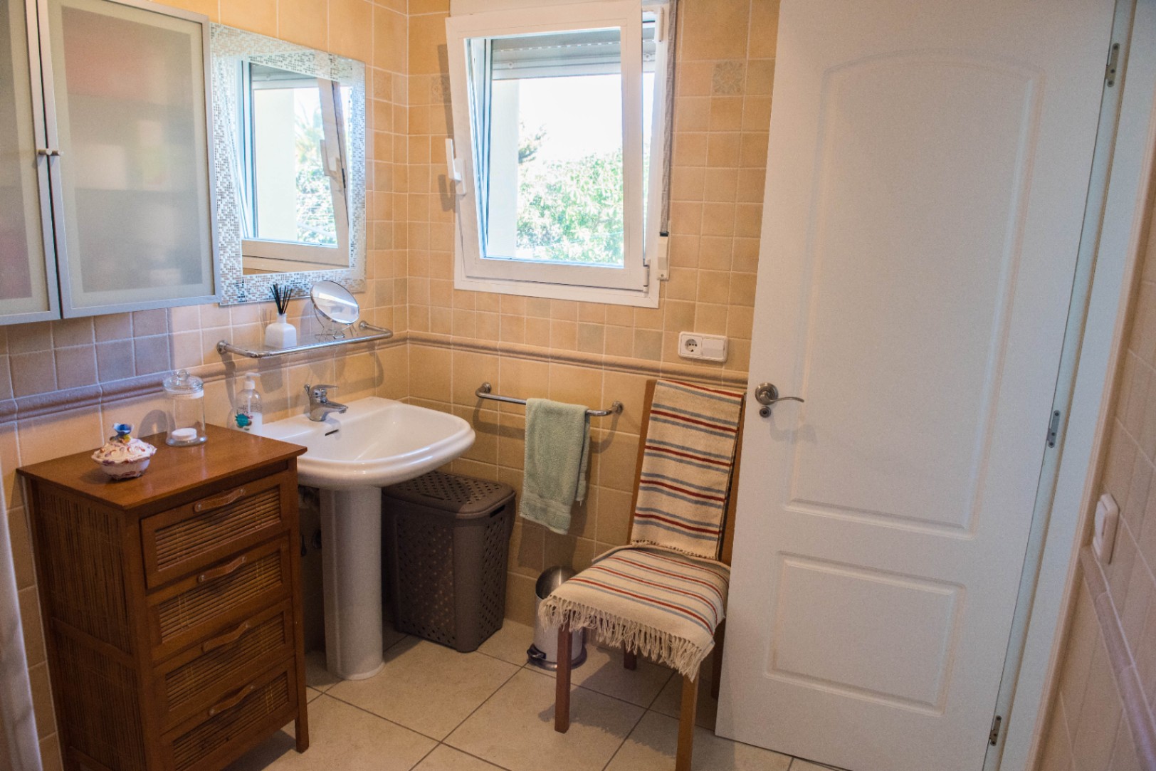 Baño con azulejos beige, lavabo de pedestal y mueble auxiliar de madera. Incluye una ventana oscilobatiente blanca que aporta luz natural.