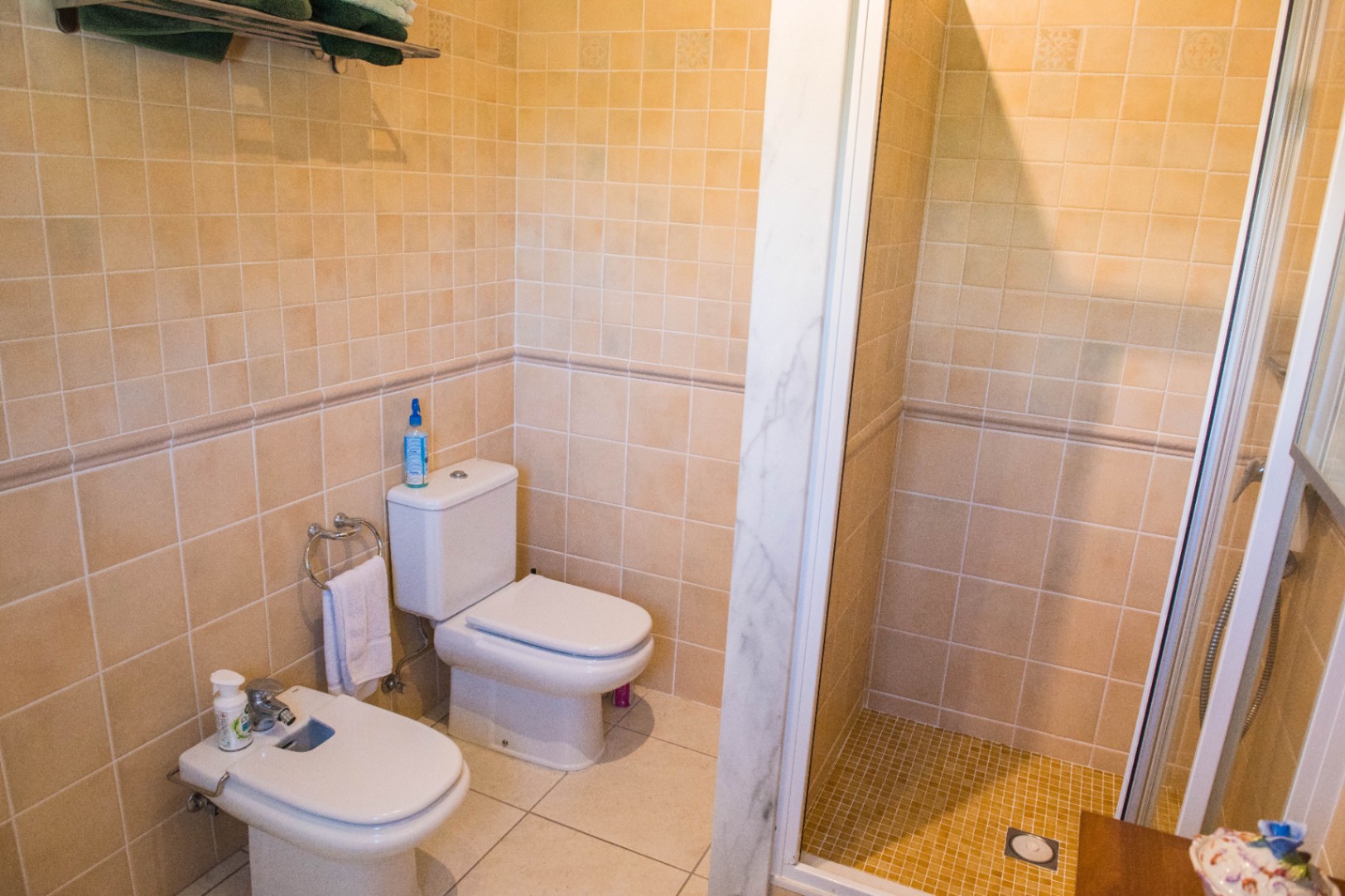 Baño con azulejos en tonos beige, inodoro y bidé de cerámica blanca, y ducha con marco de mármol y mampara de cristal transparente.