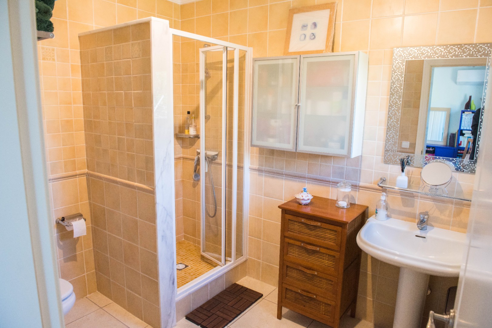 Baño con ducha de obra cerrada en cristal, azulejos en tonos beige, lavabo de pedestal blanco y un mueble auxiliar de madera con frontales trenzados.