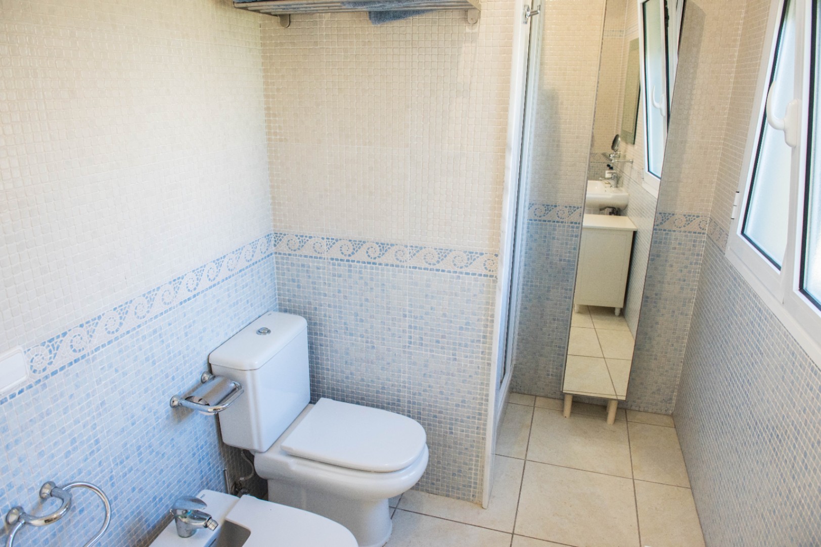 Baño con paredes de mosaico en azul y blanco, cenefa ornamental, sanitarios de cerámica blanca y ventana que aporta iluminación natural.