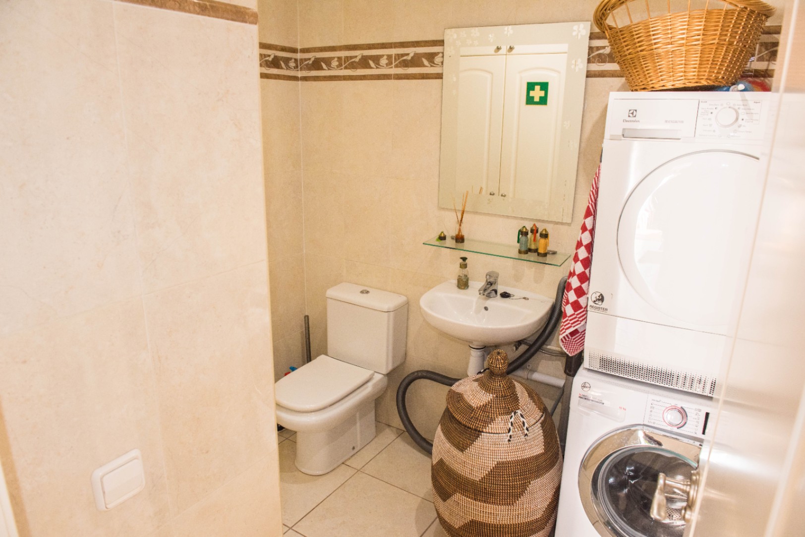 Baño funcional con zona de lavandería integrada, paredes alicatadas en beige, inodoro blanco y torre de lavado con lavadora y secadora apiladas.
