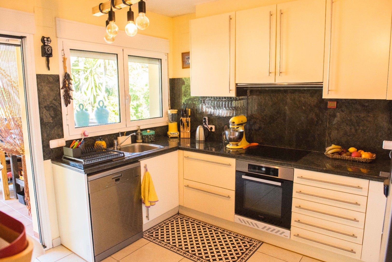 Cocina en L con armarios en tono crema, encimeras de granito oscuro y electrodomésticos integrados. El ventanal ofrece luz natural y vistas al exterior ajardinado.