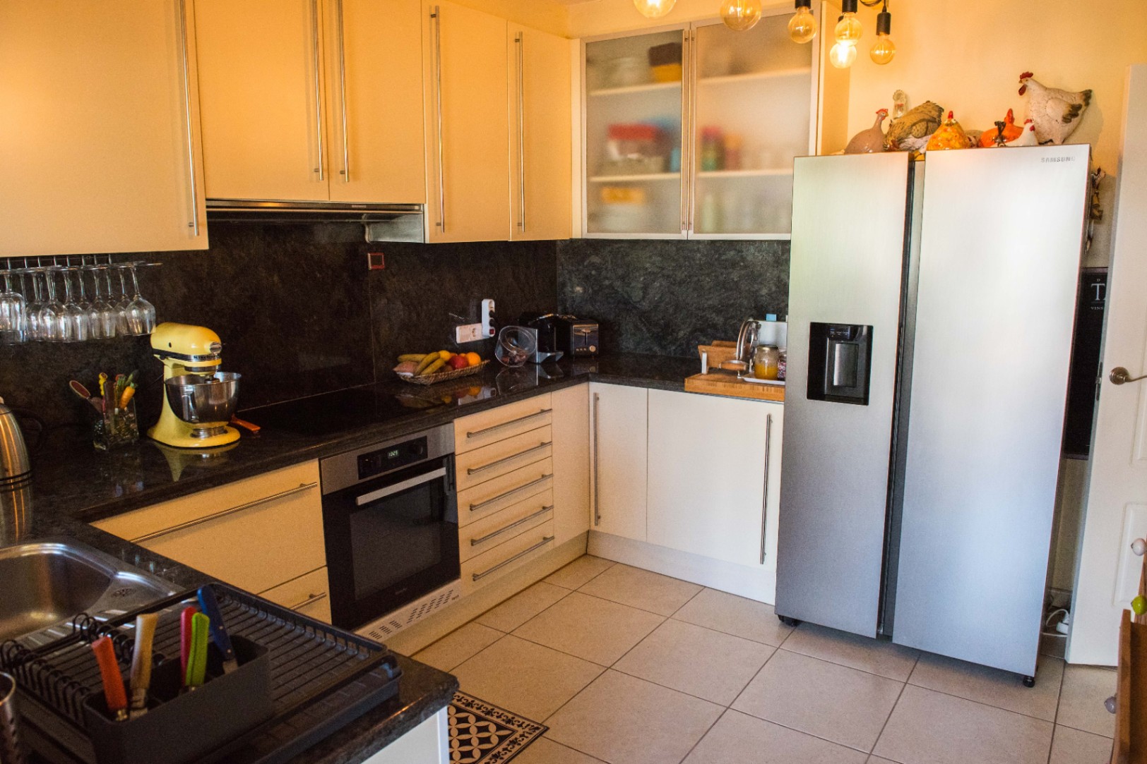 Cocina moderna en forma de L con encimeras de granito oscuro, armarios color crema y frigorífico Samsung de acero inoxidable. Incluye iluminación colgante de bombillas vistas.