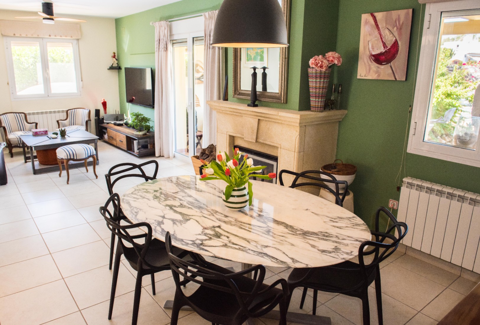 Comedor con mesa de mármol ovalada y sillas de diseño negras, junto a una chimenea de piedra y paredes verdes que conectan con el salón principal.