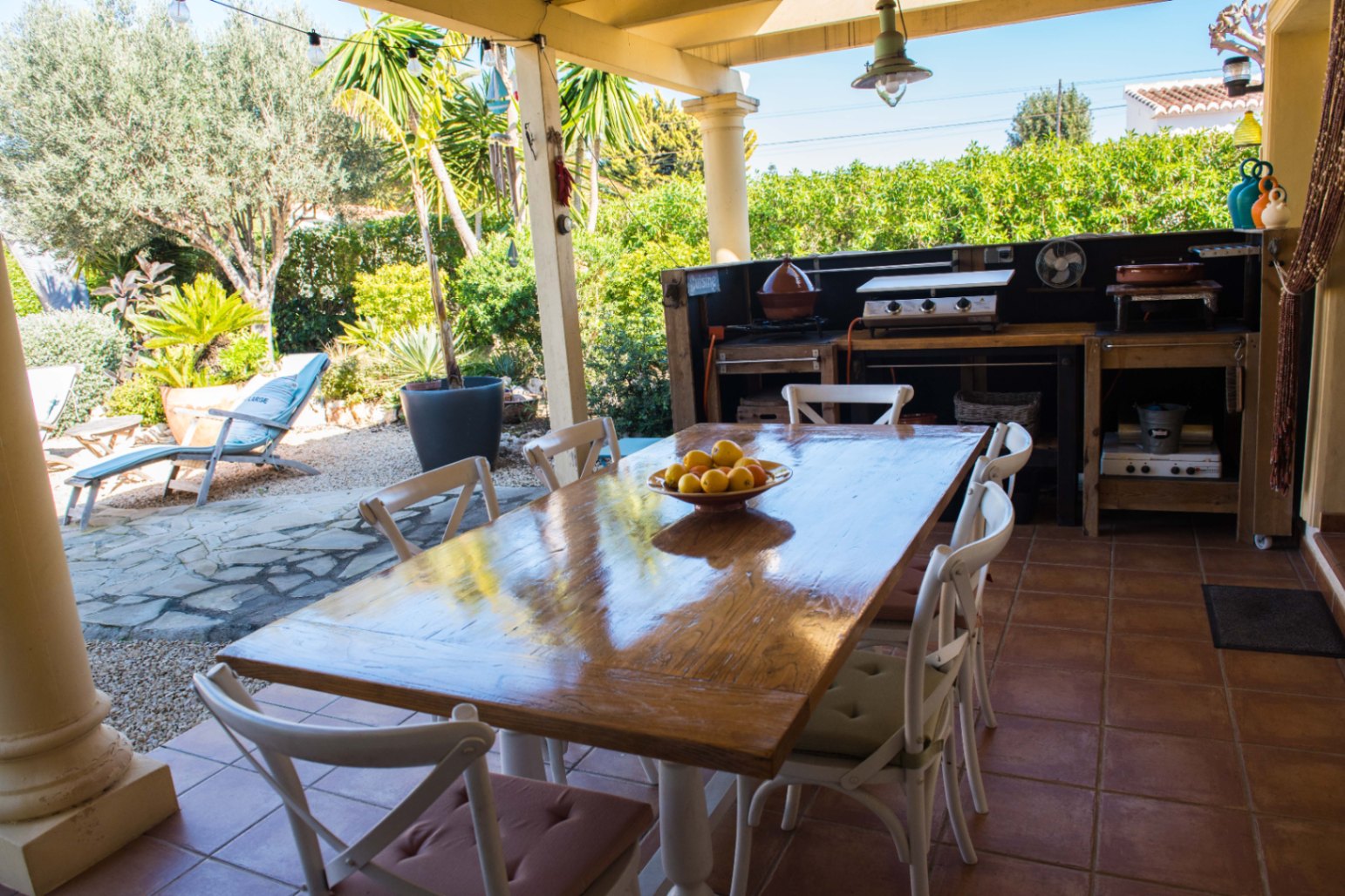 Comedor exterior en terraza de terracota con mesa de madera barnizada, cocina de verano equipada y vistas a un jardín mediterráneo con palmeras.