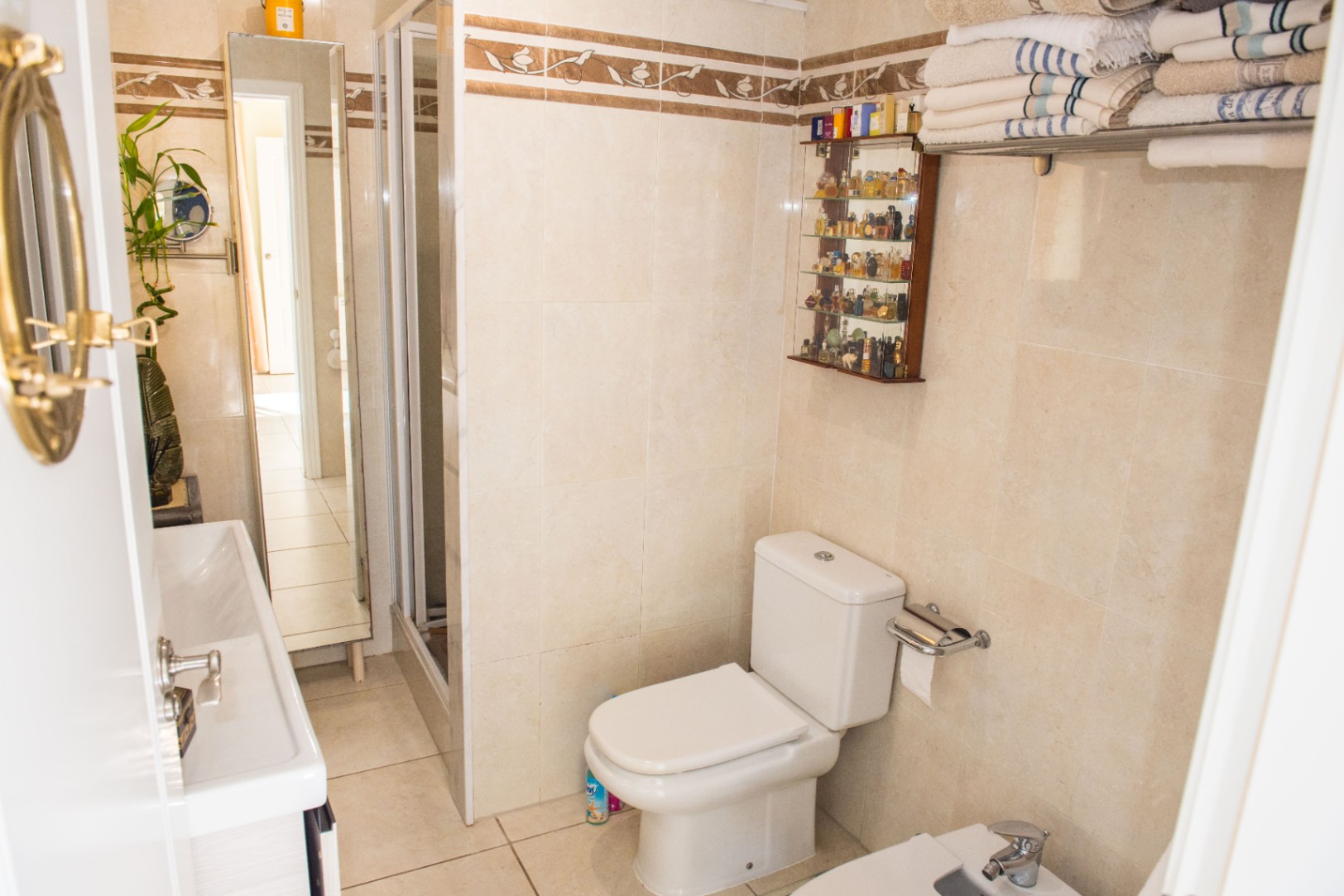 Cuarto de baño con azulejos beige y cenefa decorativa, inodoro de cerámica blanca, mampara de ducha de cristal y vitrina de pared para perfumes.