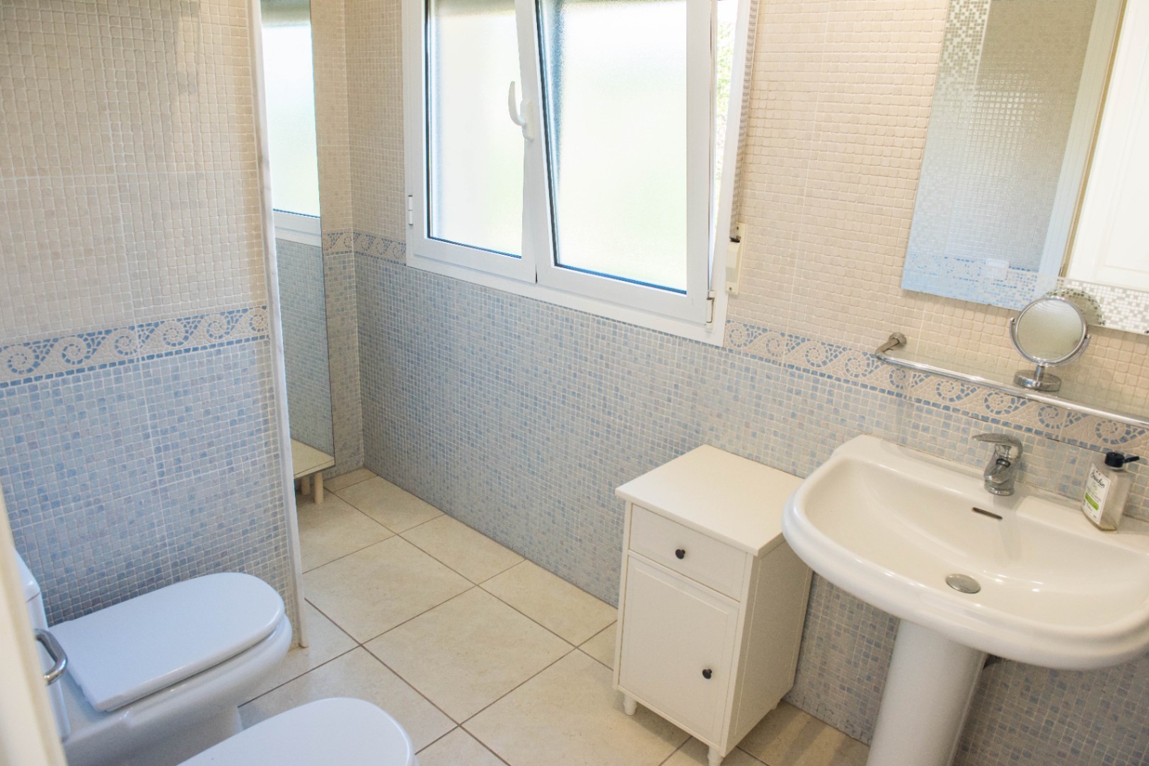 Cuarto de baño con paredes de mosaico azul y beige, lavabo de pedestal blanco y una ventana de PVC con cristal traslúcido que aporta luz natural.