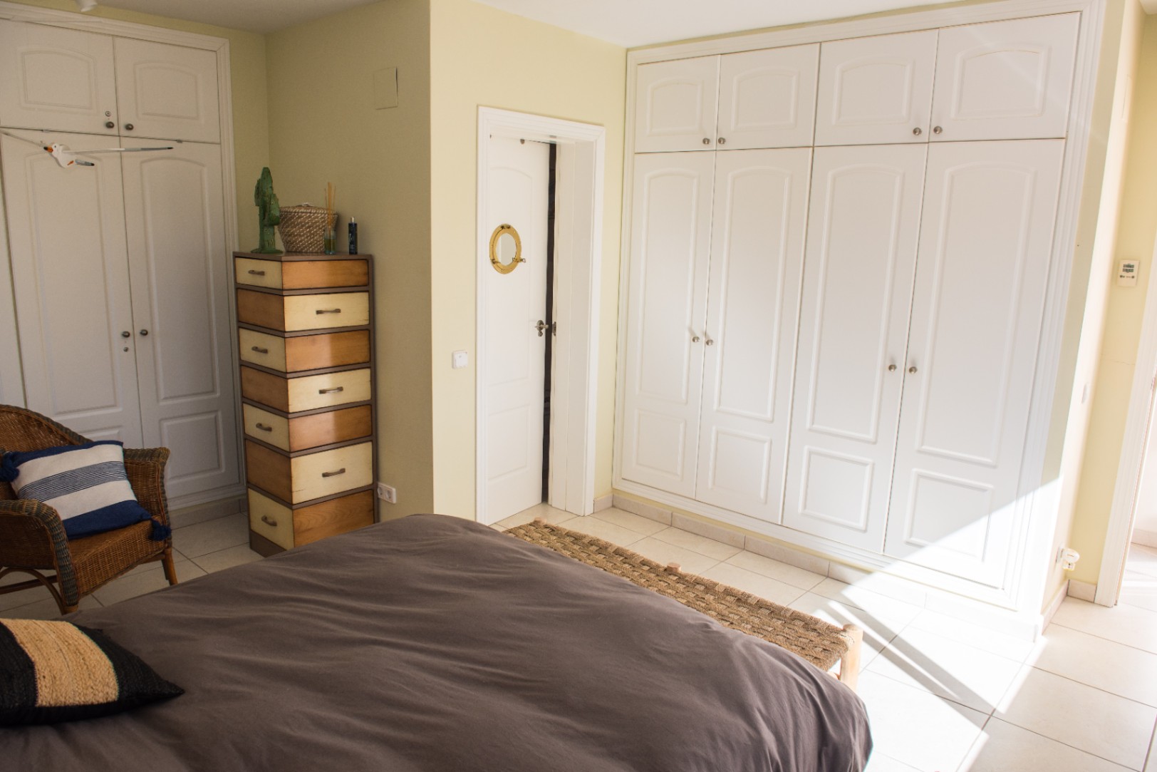 Dormitorio con amplios armarios empotrados blancos, una cómoda de madera bitono y un sillón de mimbre. La luz natural destaca el suelo cerámico y las paredes de tono neutro.