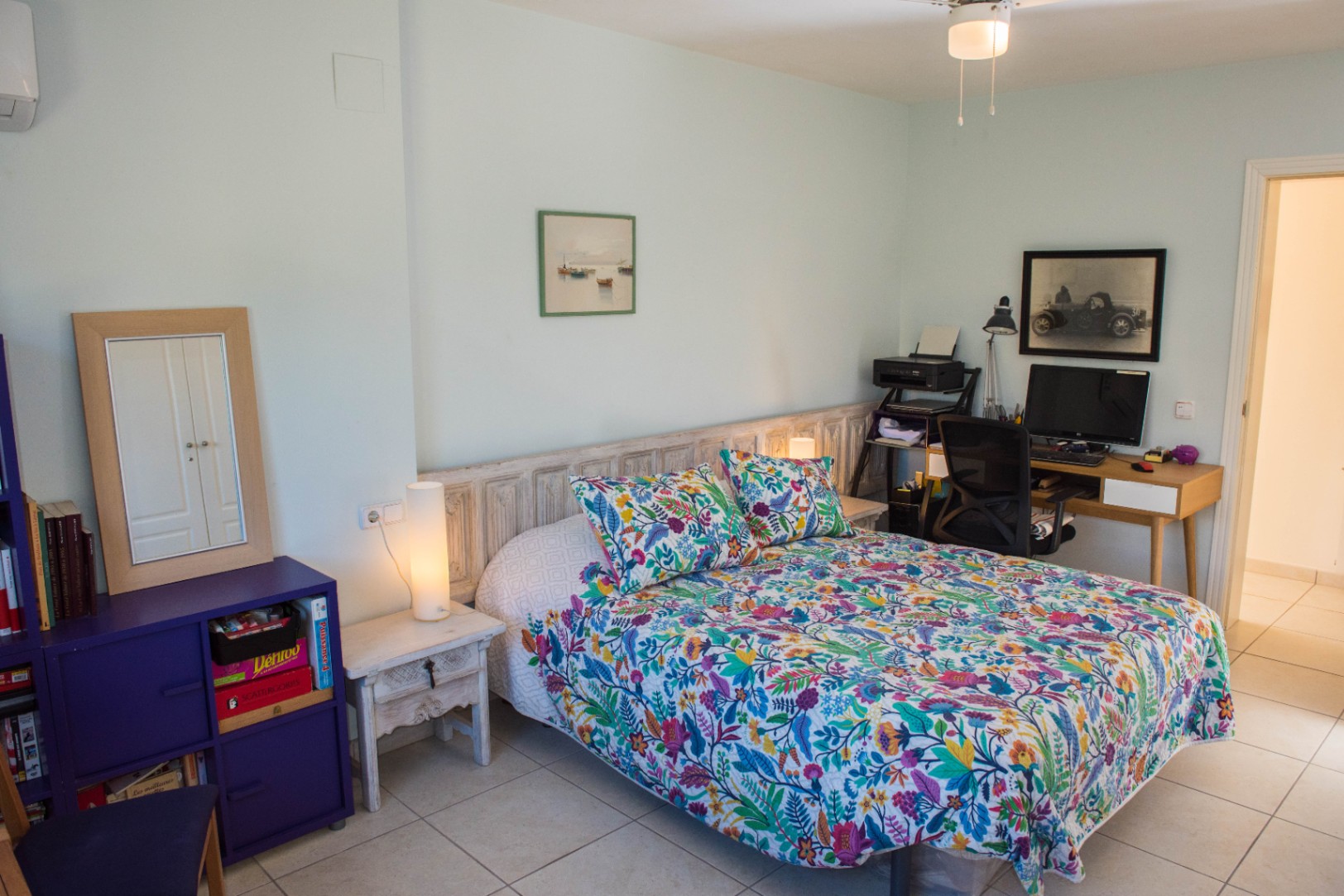 Dormitorio con cama de estampado floral, cabecero de madera rústica, zona de escritorio integrada y suelos de baldosa clara bajo iluminación ambiental.