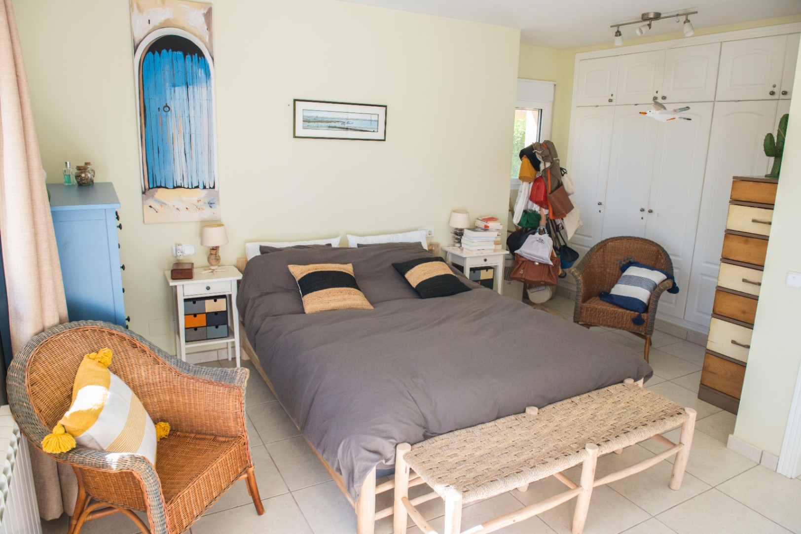 Dormitorio con cama doble, sillones de mimbre y armarios empotrados blancos. Incluye un banco de fibras naturales y suelo de baldosas claras.