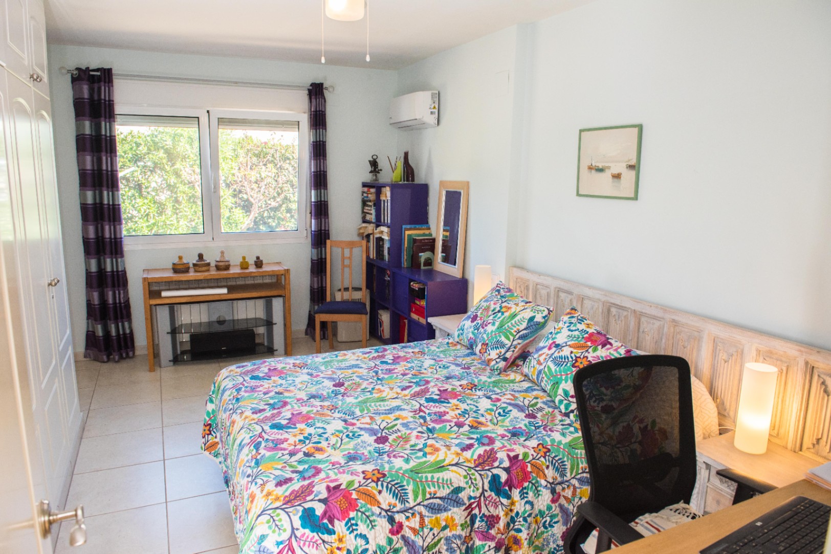 Dormitorio con colcha floral, cabecero de madera tallada y ventanal con vistas al jardín. Incluye armario empotrado, estantería púrpura y zona de escritorio.