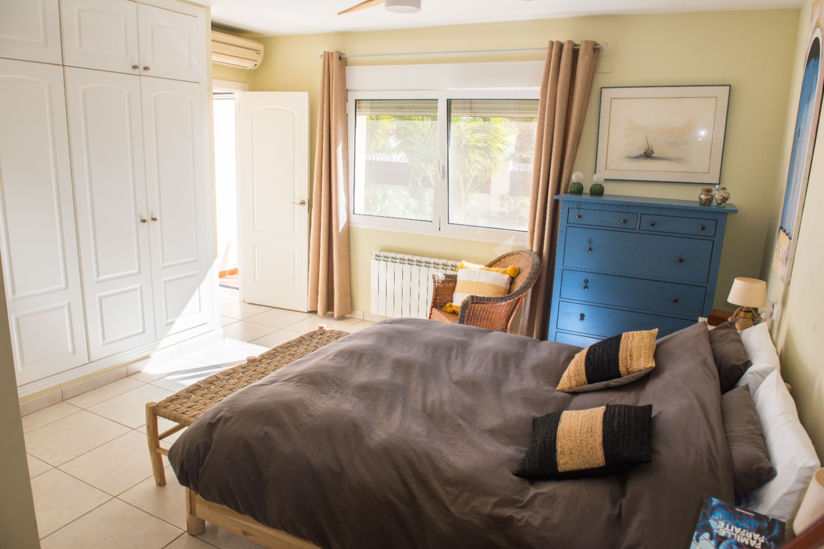 Dormitorio luminoso con armarios empotrados blancos, cómoda de madera azul y suelos de baldosa. Incluye ventanal con vistas al jardín y aire acondicionado tipo split.