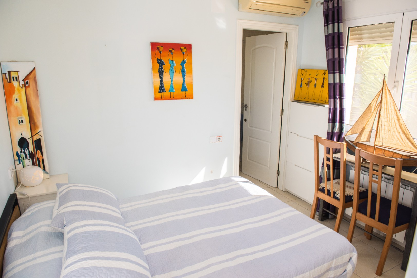 Dormitorio luminoso con ropa de cama de rayas azules, arte decorativo y un rincón de trabajo con sillas de madera y un velero ornamental junto a la ventana.