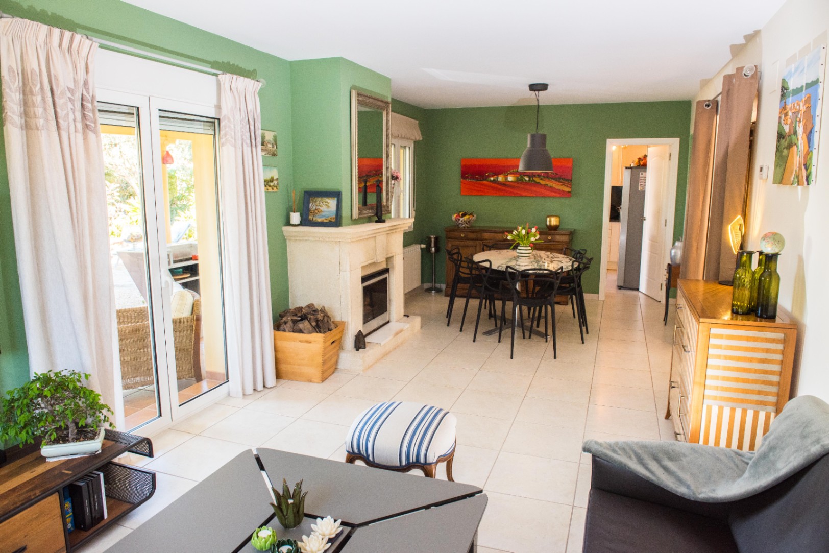 Salón-comedor con paredes en verde salvia, chimenea de piedra natural y suelos cerámicos claros. Grandes ventanales correderos conectan el interior con la terraza exterior.