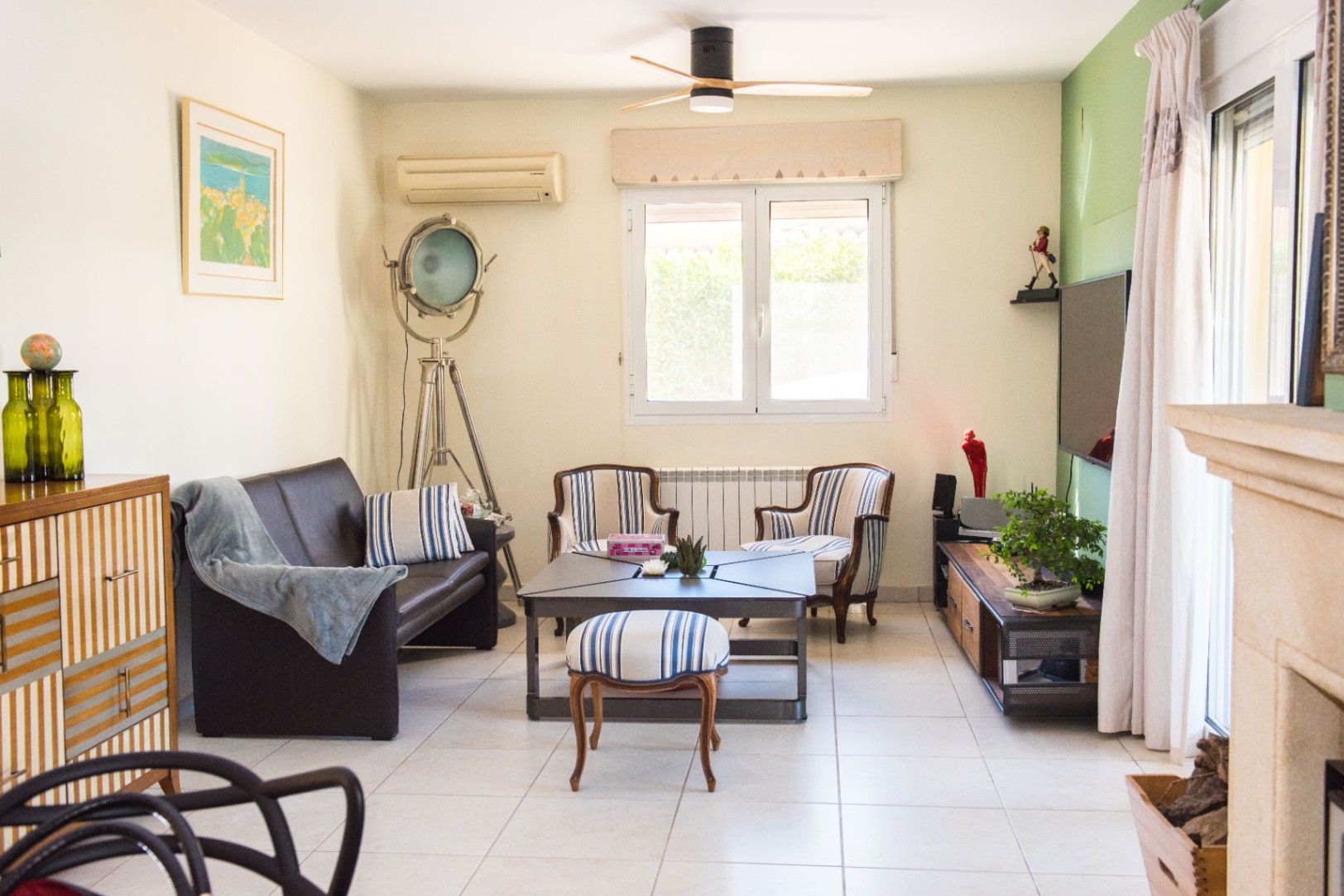Salón luminoso con sofá de cuero marrón, sillones de rayas y una lámpara de foco industrial. Cuenta con ventilador de techo, suelo cerámico y una pared de acento verde.