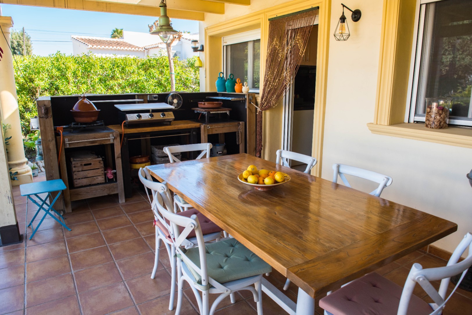 Terraza cubierta con amplia mesa de madera, sillas blancas y cocina exterior equipada con plancha y detalles rústicos sobre suelo de terracota.