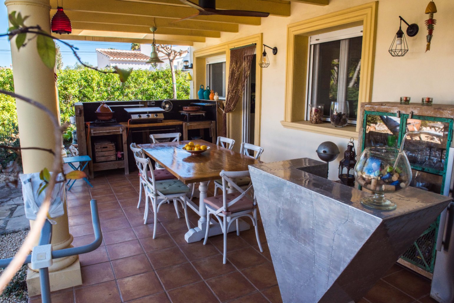 Terraza cubierta con cocina exterior rústica, mesa de comedor de madera y una barra metálica industrial sobre suelos de baldosas de terracota.