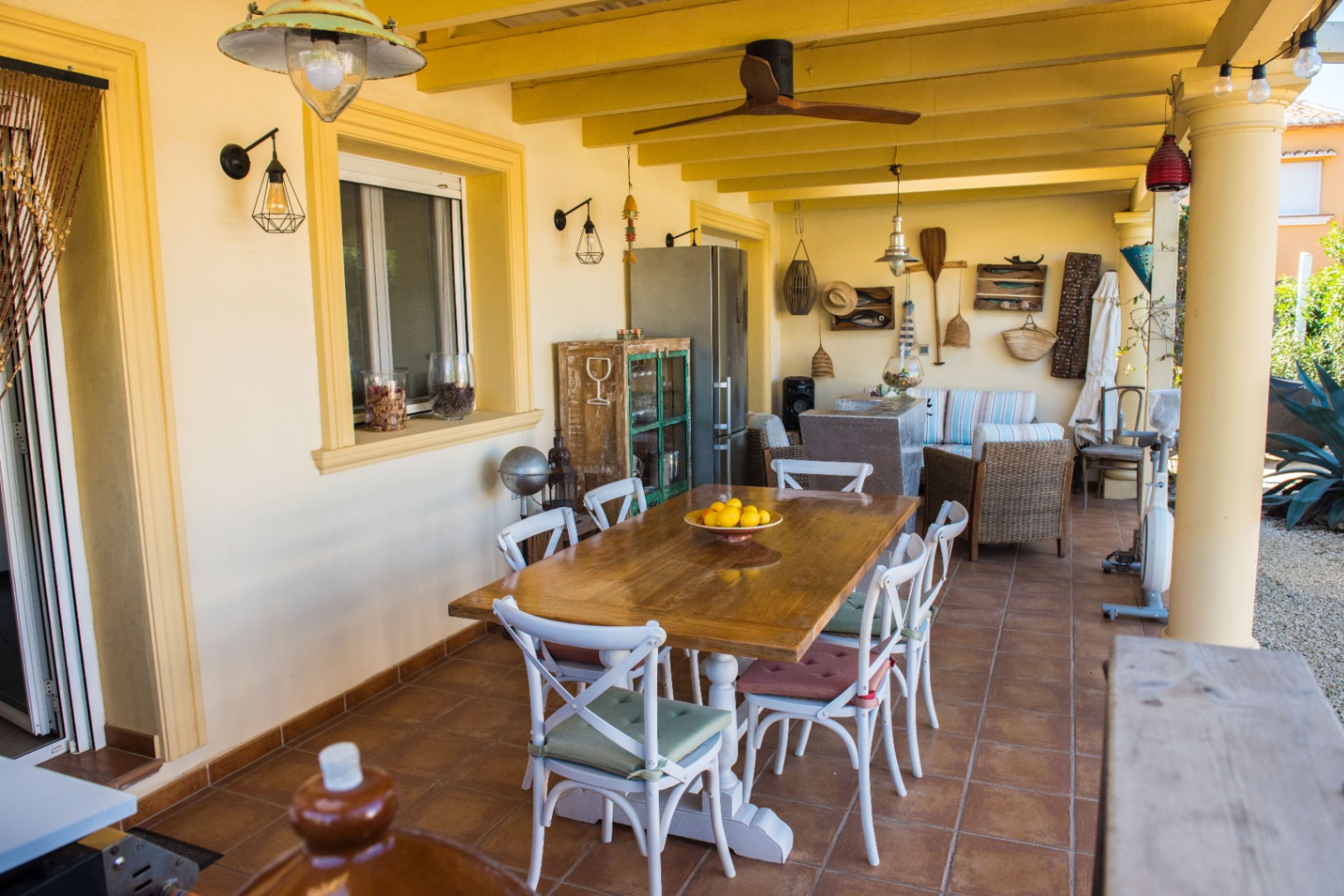 Terraza cubierta con mesa de comedor de madera, sillas blancas de aspa, decoración rústica y techo de vigas amarillas con ventilador.