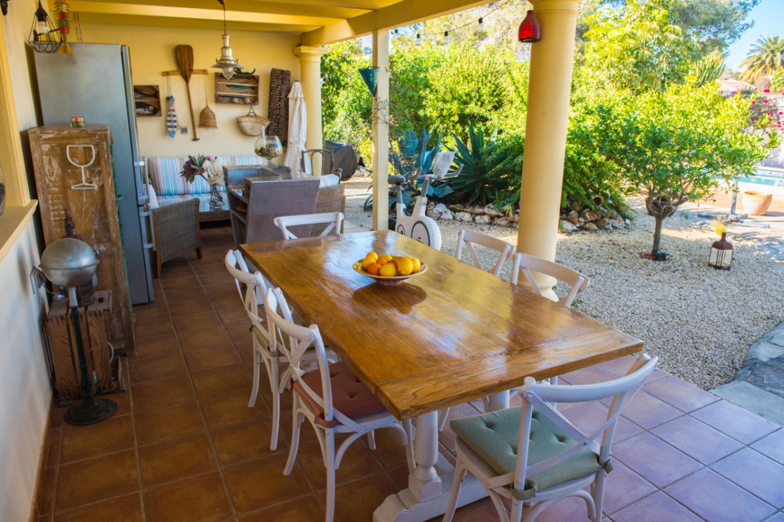 Terraza cubierta con mesa de madera rústica, sillas blancas y suelo de terracota, con vistas a un jardín mediterráneo de grava y vegetación autóctona.