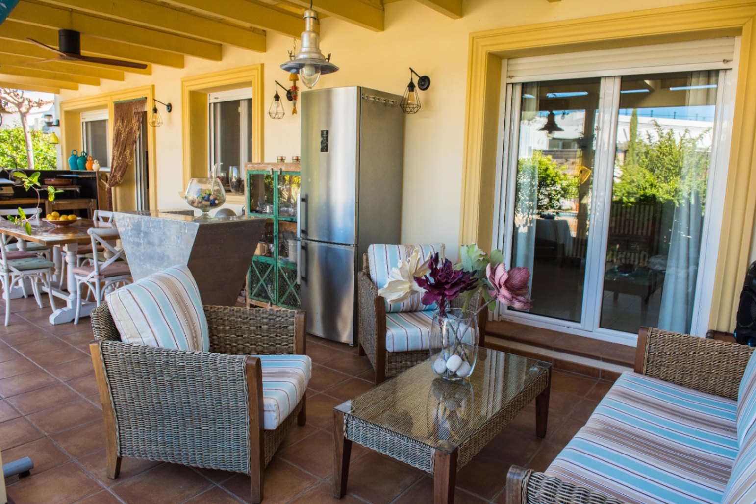 Terraza cubierta con mobiliario de mimbre, cojines de rayas, suelo de terracota y zona de cocina exterior equipada con frigorífico de acero inoxidable.