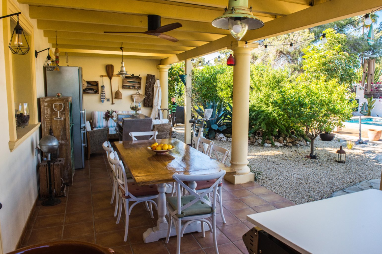 Terraza cubierta con suelo de terracota, mesa de comedor de madera y zona de estar rústica con vistas al jardín mediterráneo y la piscina.