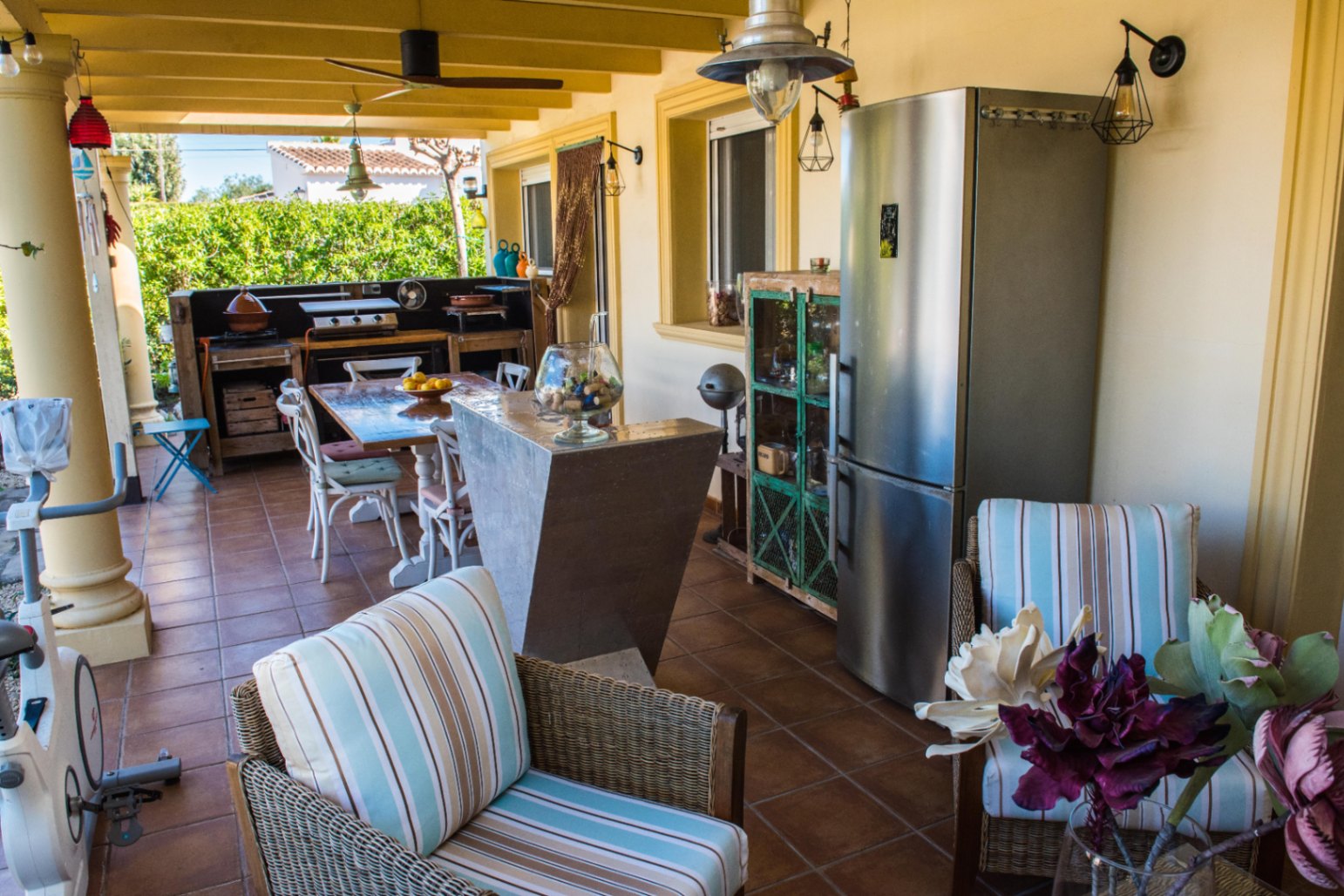 Terraza exterior cubierta con cocina rústica, frigorífico de acero inoxidable y zona de comedor sobre suelos de terracota y vigas vistas.