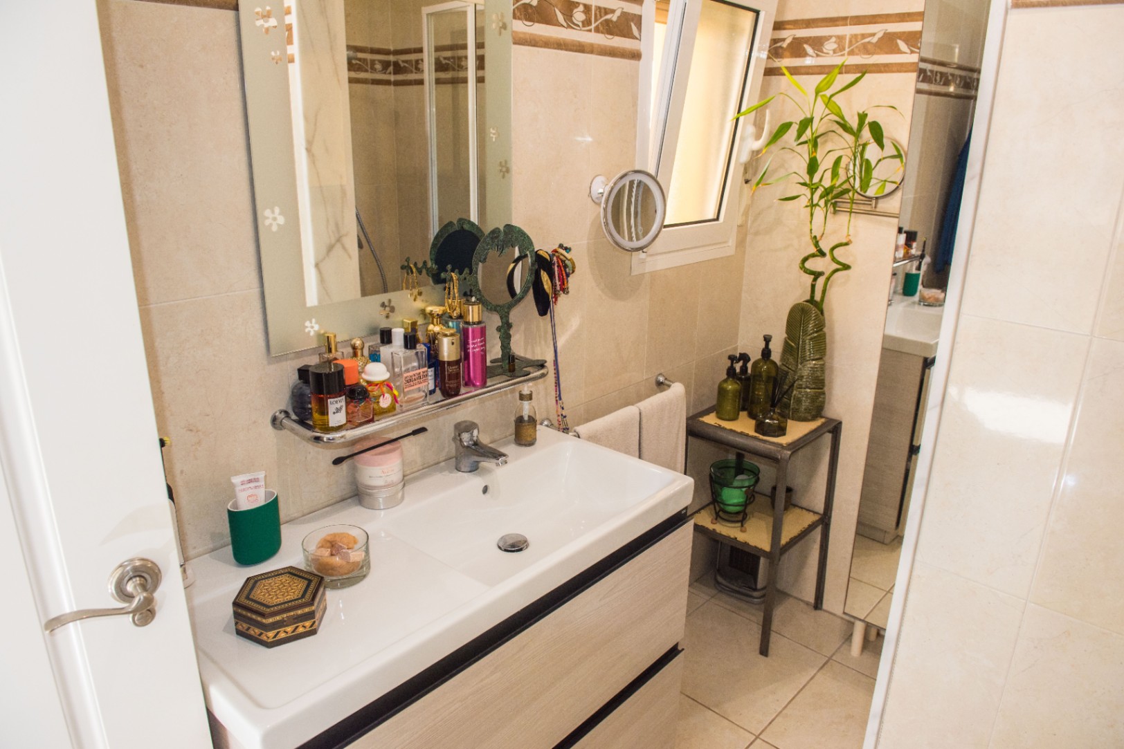 Tocador de baño con lavabo de cerámica blanca, mueble de madera clara y espejo decorado. Incluye estantería auxiliar, luz natural lateral y revestimiento cerámico en tonos crema.