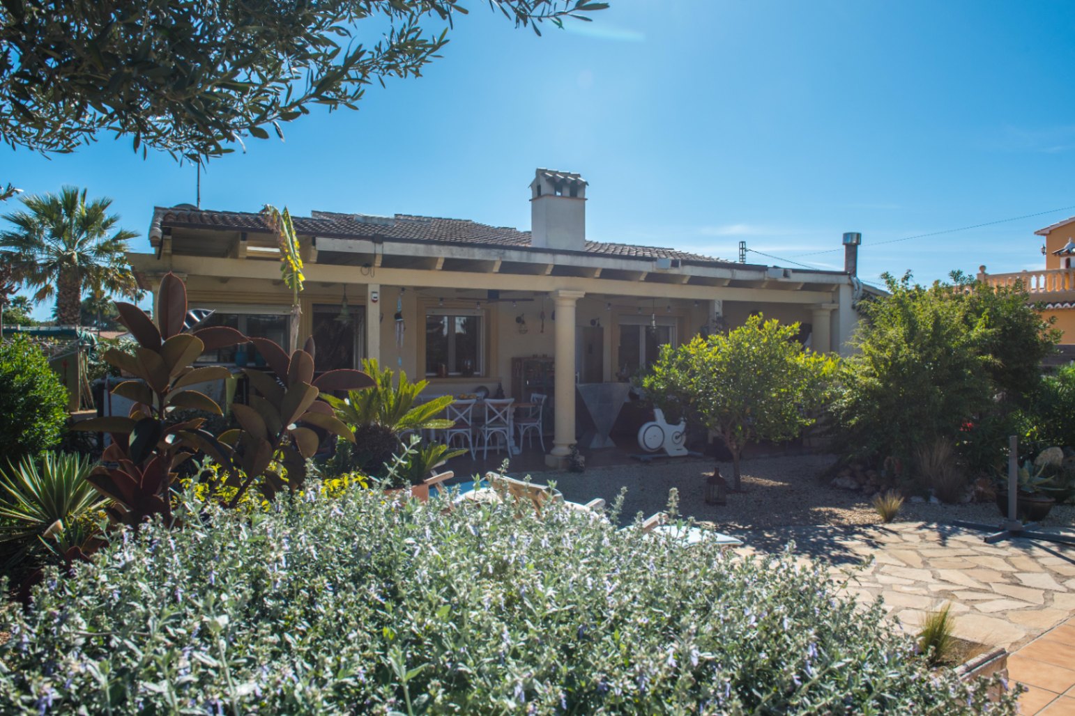 Vista exterior de una villa con porche cubierto, vigas de madera y jardín mediterráneo con senderos de piedra y vegetación autóctona bajo el sol.