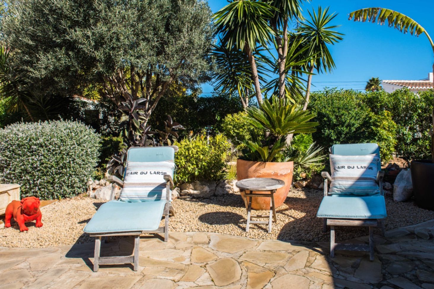 Zona de descanso exterior con tumbonas de madera, cojines azules y jardín mediterráneo con olivos, palmeras y suelo de piedra natural y grava.