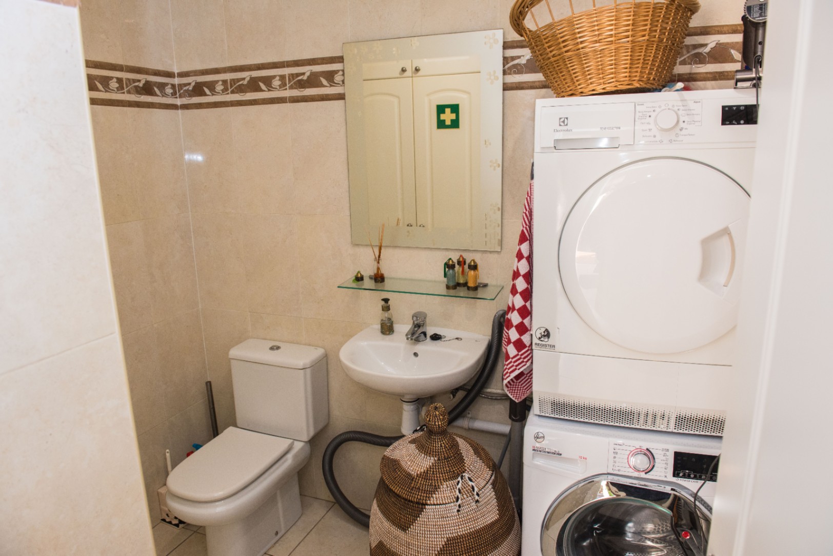 Zona funcional de lavandería y baño con electrodomésticos apilados, inodoro cerámico y paredes de azulejos beige con cenefa decorativa.