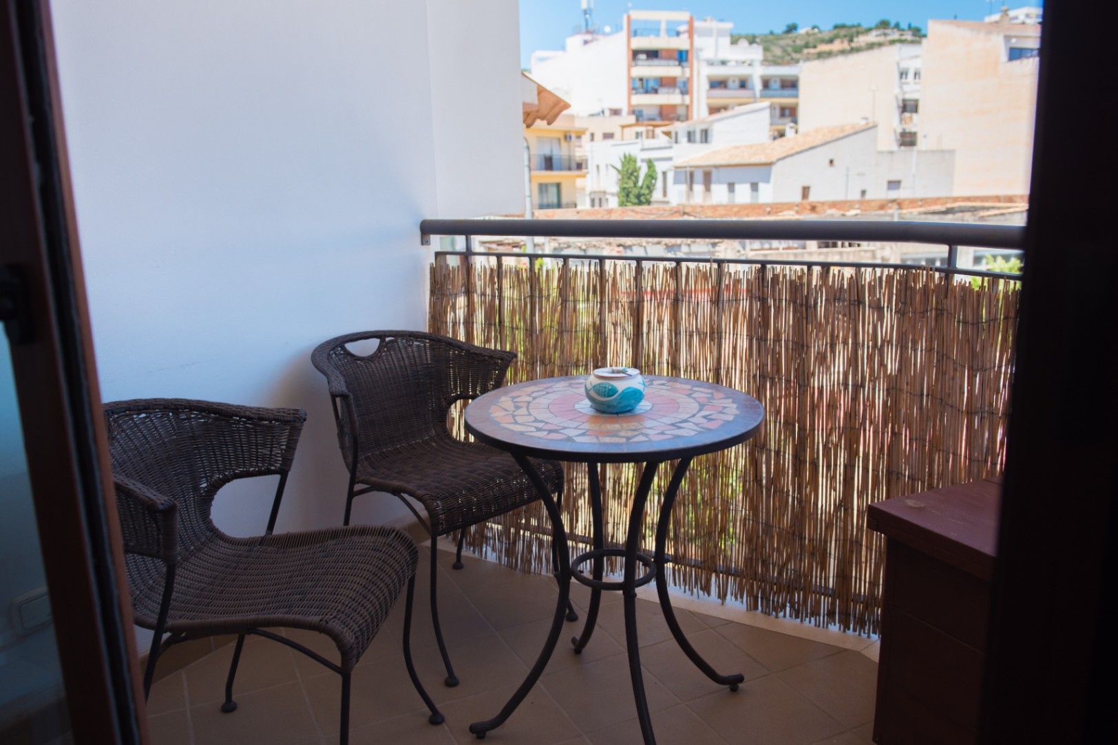 Balcón privado con mesa de mosaico, dos sillas de mimbre y pantalla de cañizo, con vistas despejadas al entorno urbano residencial.