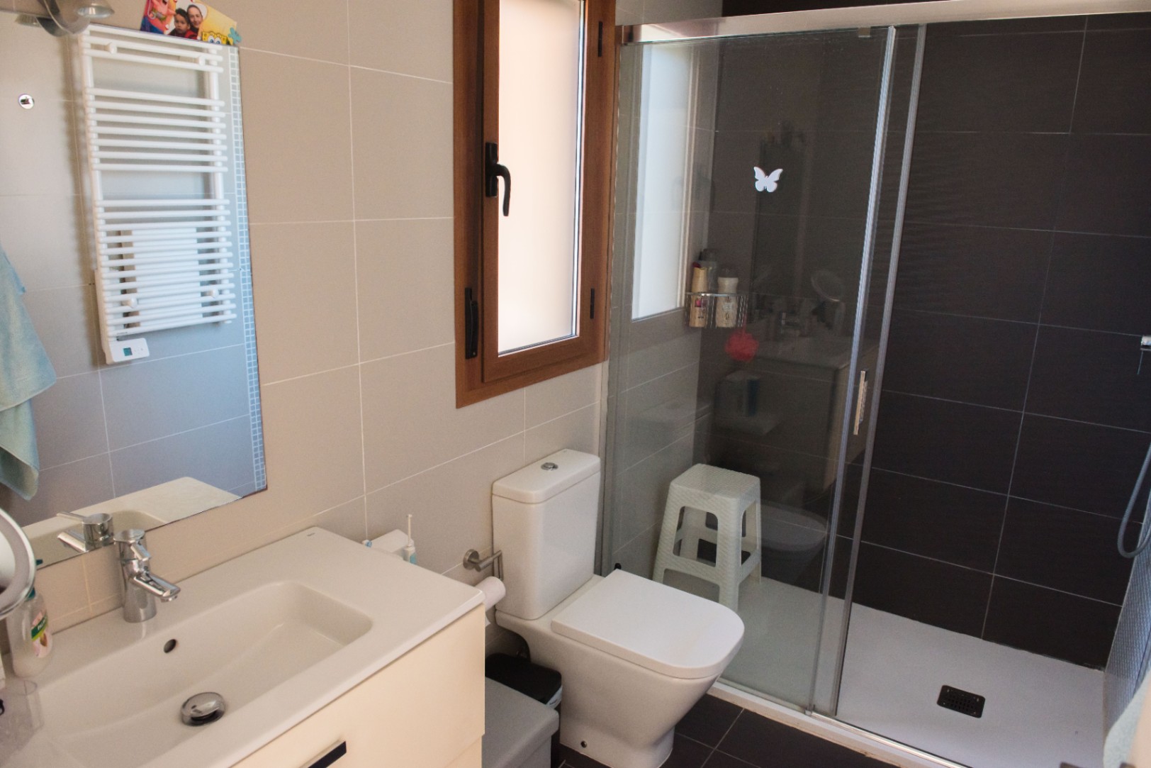 Baño funcional con ducha de obra en tonos oscuros, lavabo blanco con mueble y radiador toallero. Dispone de una ventana con carpintería de madera.