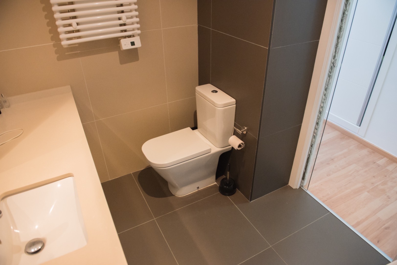 Baño moderno con inodoro de cerámica blanca, suelo de baldosas gris oscuro y radiador toallero. Incluye un lavabo integrado en encimera de tono crema.