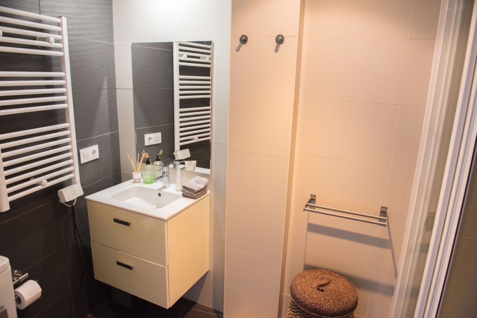 Baño moderno con mueble suspendido de dos cajones, radiador toallero blanco y revestimiento cerámico en tonos gris oscuro y crema.