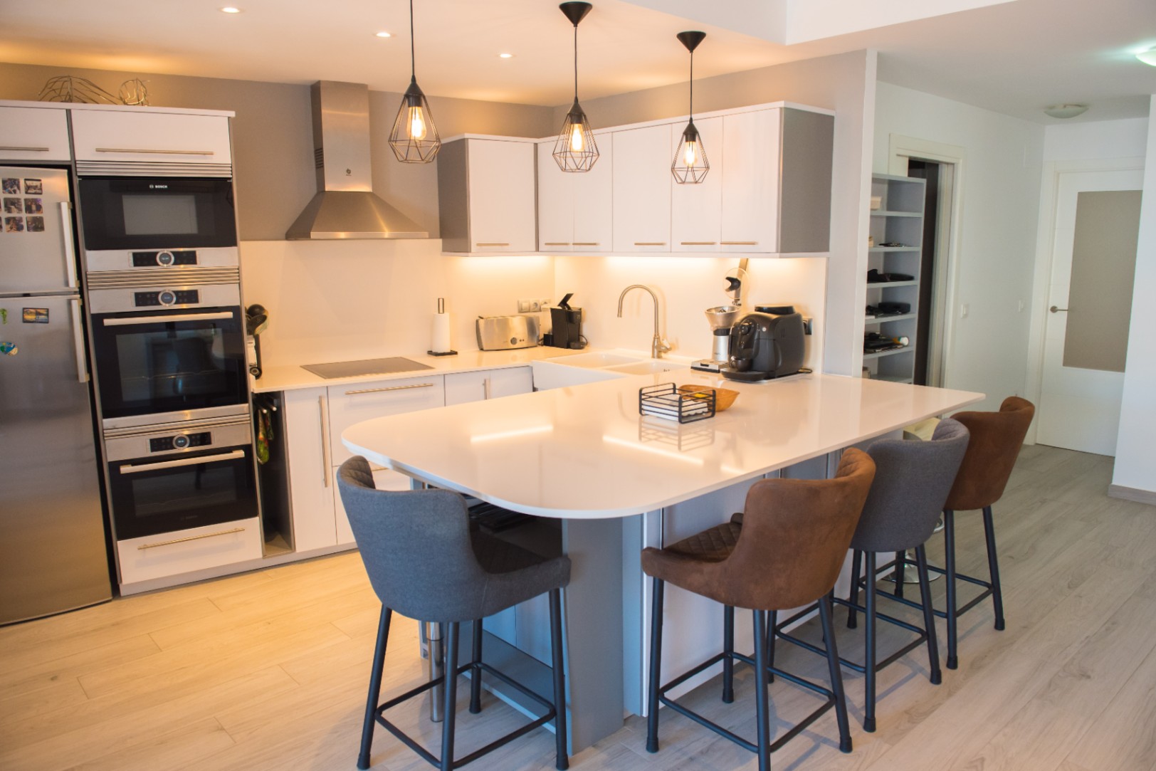 Cocina moderna con isla blanca y taburetes, electrodomésticos Bosch integrados, iluminación colgante industrial y suelos de madera clara.