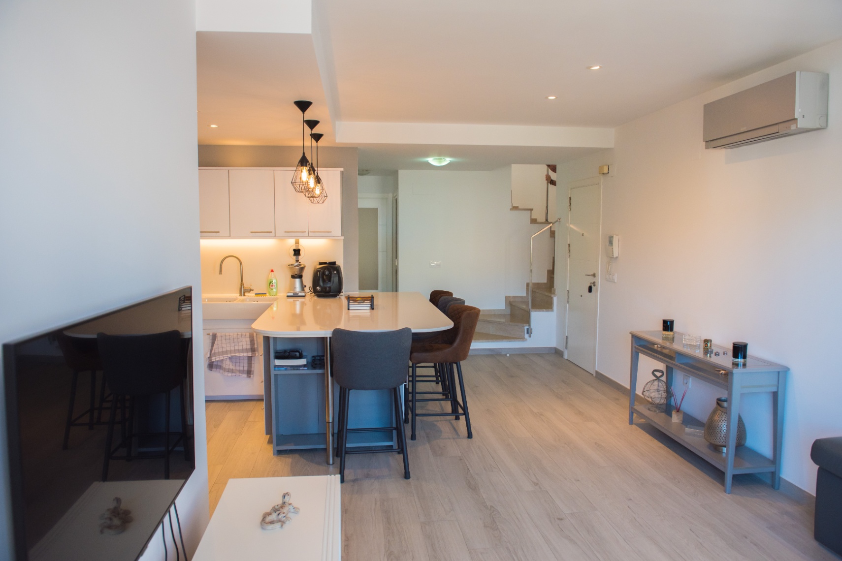 Cocina moderna de concepto abierto con una isla blanca, taburetes tapizados, lámparas colgantes industriales y suelos de madera clara junto a una escalera.