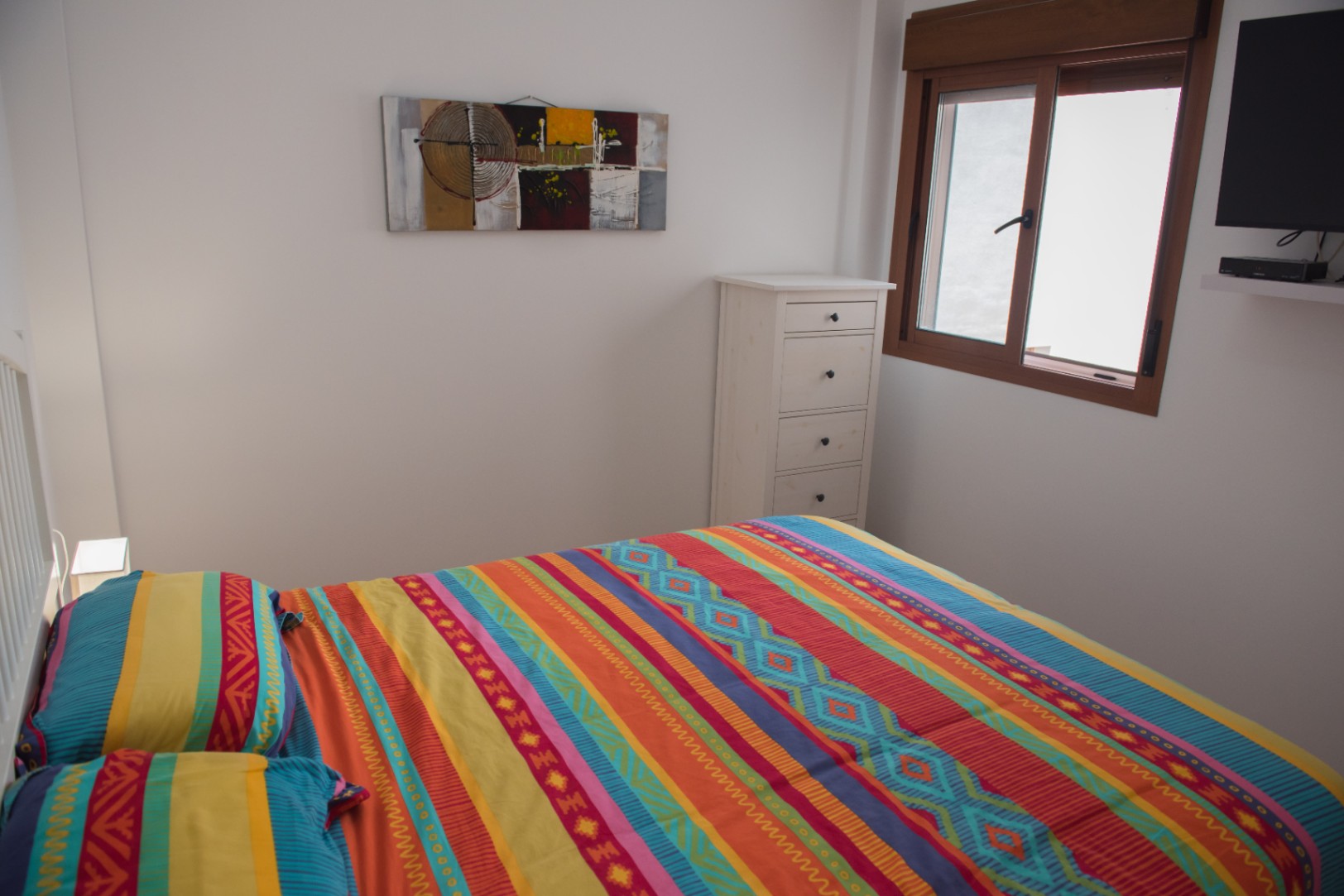 Dormitorio con cama de textiles coloridos, cómoda blanca de madera y televisión de pared junto a una ventana con marco de madera.