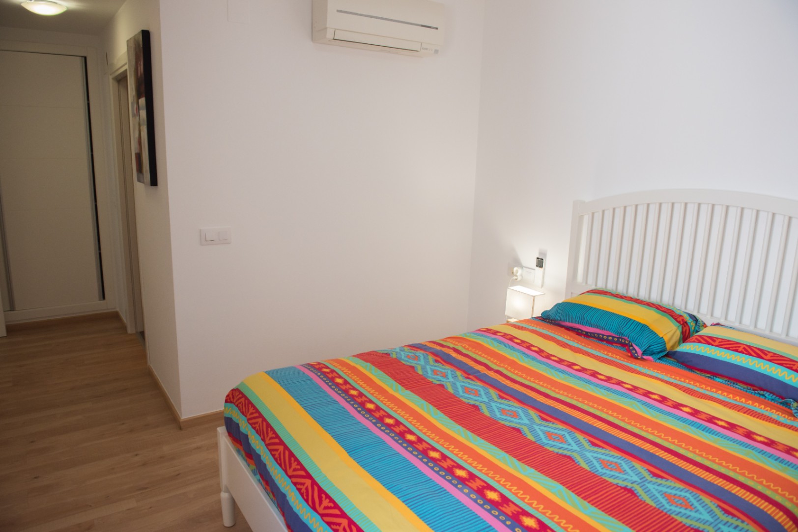 Dormitorio luminoso con cama de estructura blanca, ropa de cama estampada y suelos de madera clara. Incluye aire acondicionado split y paredes blancas.