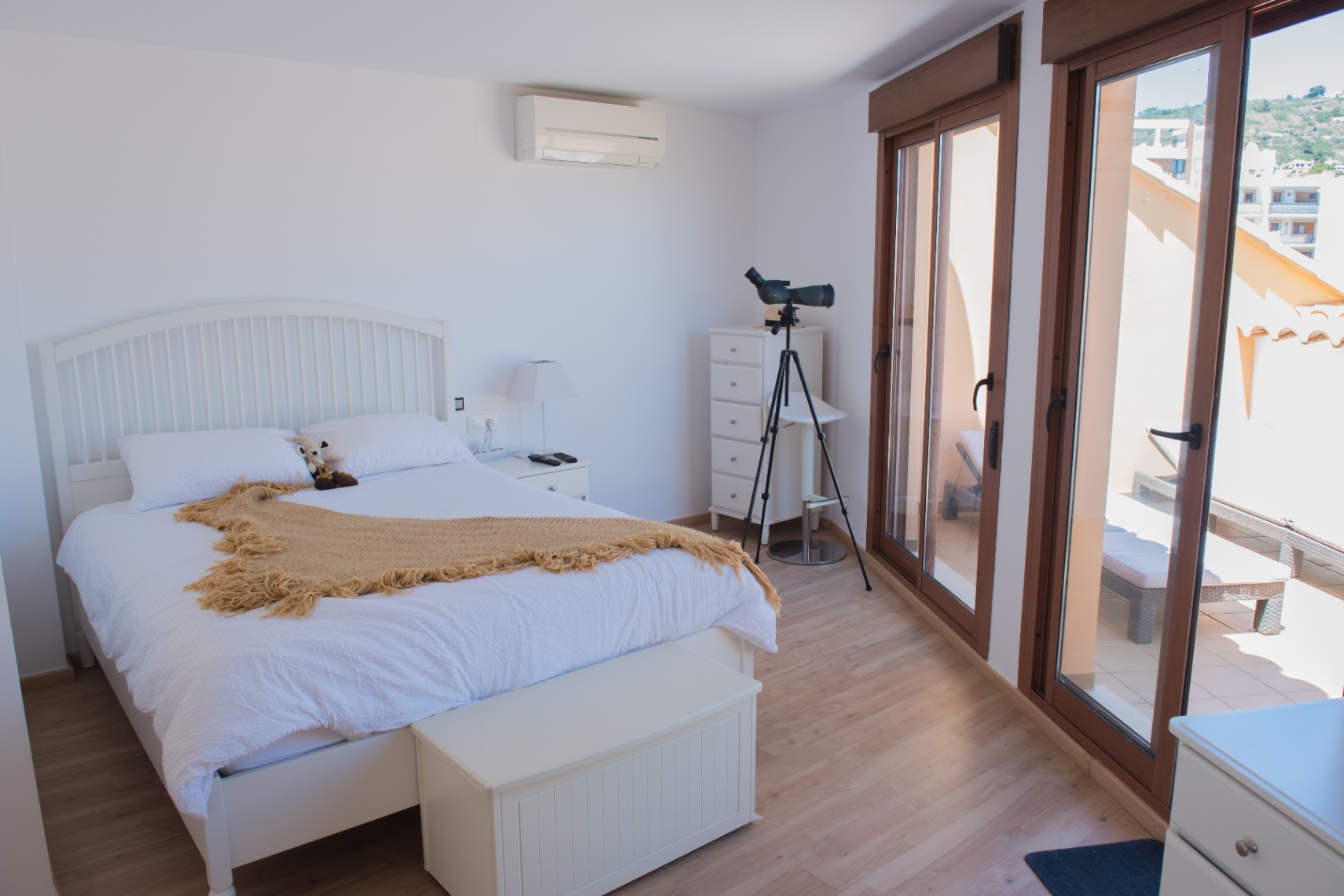 Dormitorio luminoso con mobiliario blanco, suelos de madera clara y telescopio. Grandes puertas correderas dan acceso a una terraza privada con tumbonas y vistas.