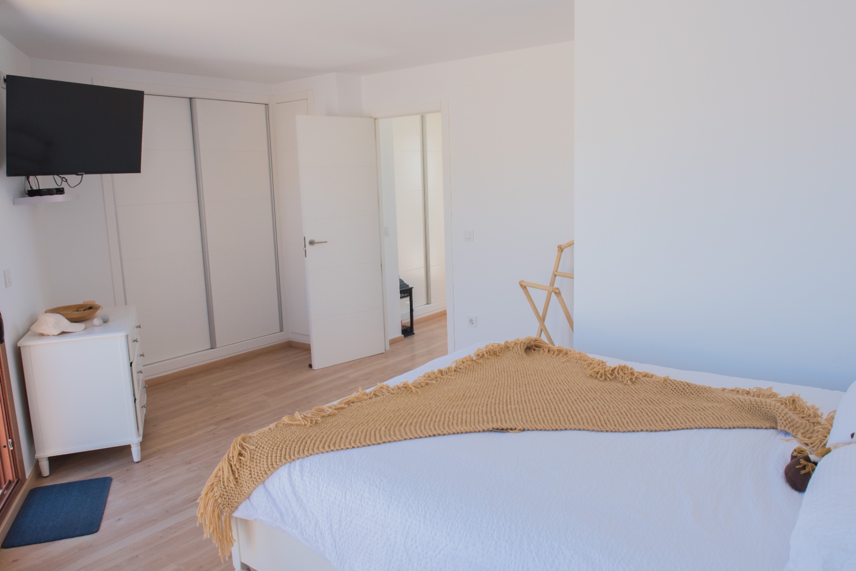 Dormitorio moderno con suelos de madera clara, armario empotrado de puertas correderas y televisión de pared. Incluye una cómoda blanca y decoración minimalista en tonos neutros.