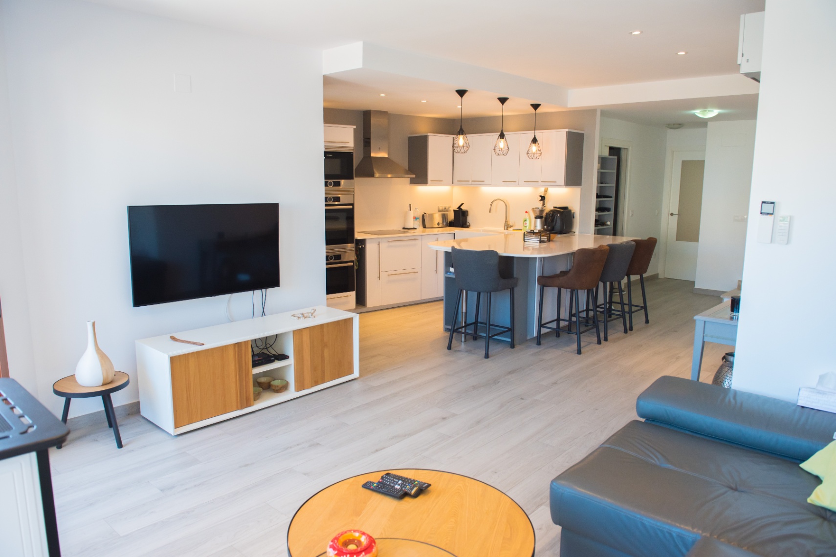 Espacio diáfano que integra una cocina moderna con isla blanca, electrodomésticos encastrados y un salón con suelos de tarima clara y mobiliario funcional.
