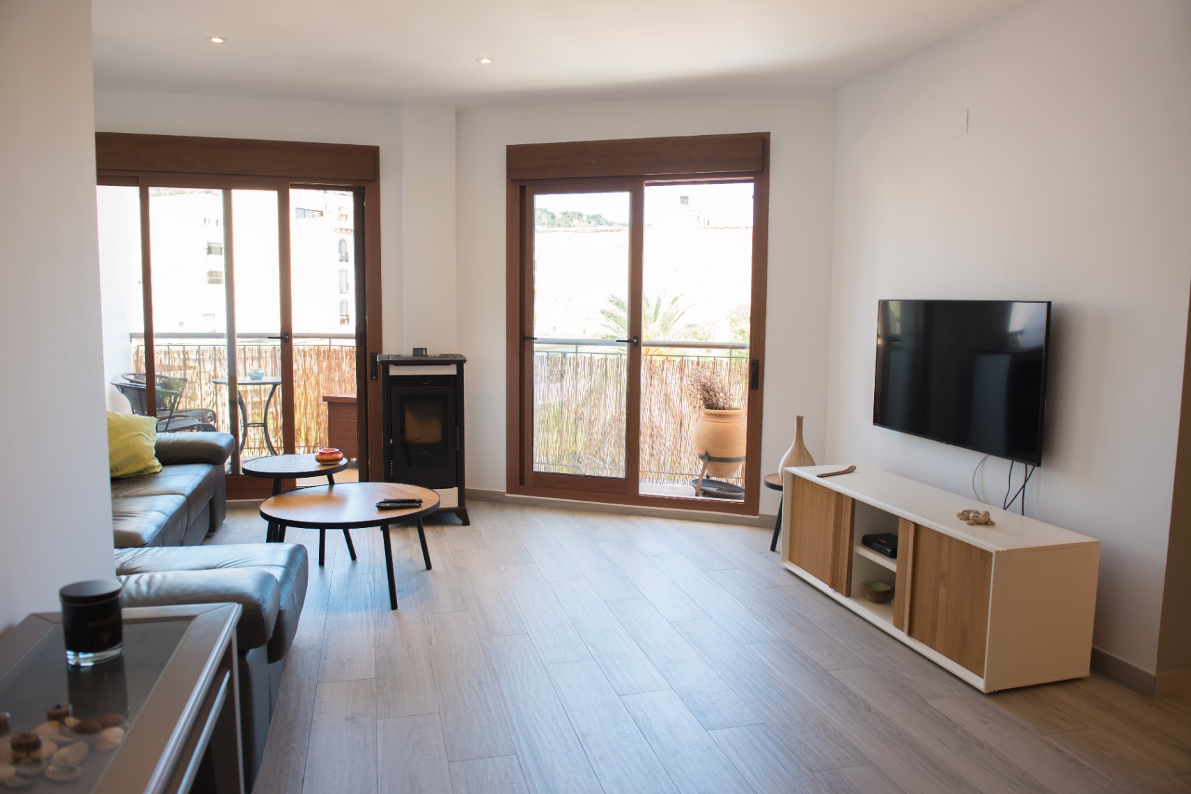 Salón contemporáneo con sofá de cuero gris, estufa de pellets y mesas de madera. Amplios ventanales con marco de madera conectan con el balcón exterior.