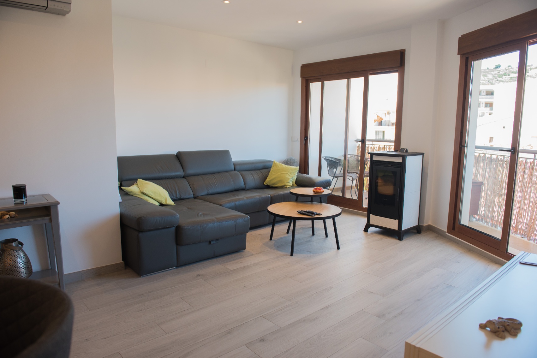 Salón moderno con sofá rinconera de cuero oscuro, mesas de centro circulares de madera y estufa de pellets. Grandes ventanales de madera ofrecen luz natural y acceso al balcón.