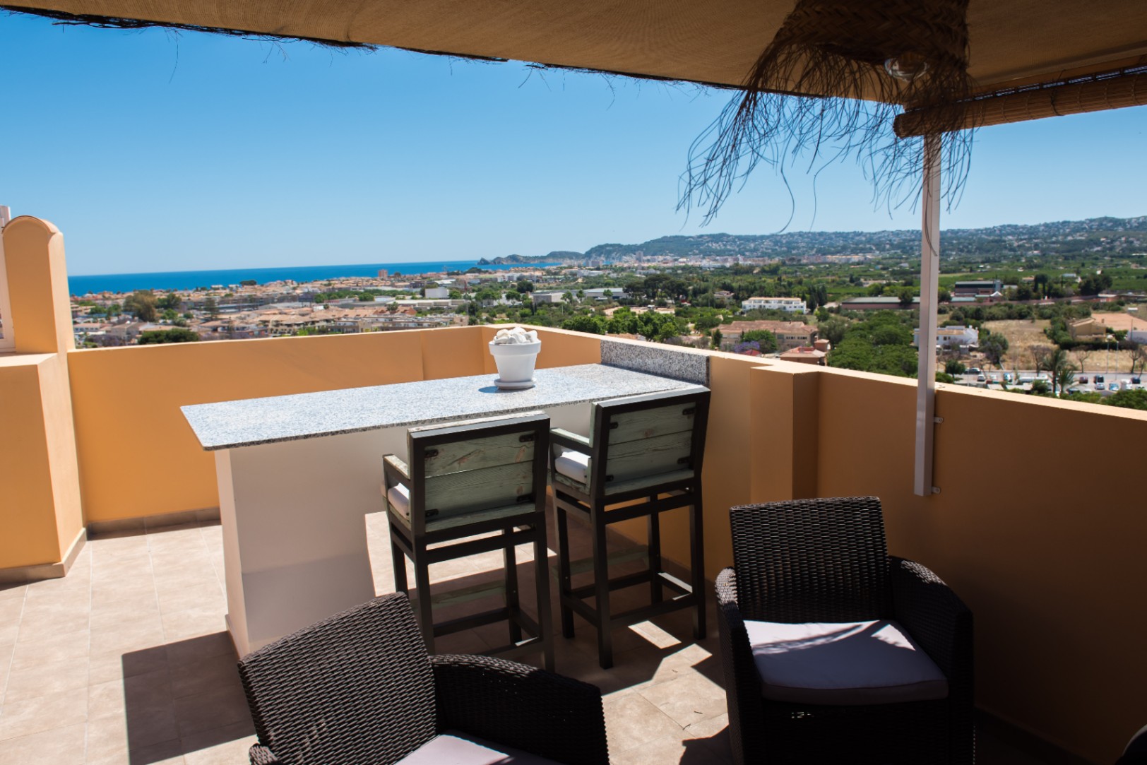 Terraza con mesa de bar de granito, taburetes altos y sillones de mimbre, que ofrece vistas panorámicas del pueblo costero y el mar Mediterráneo.