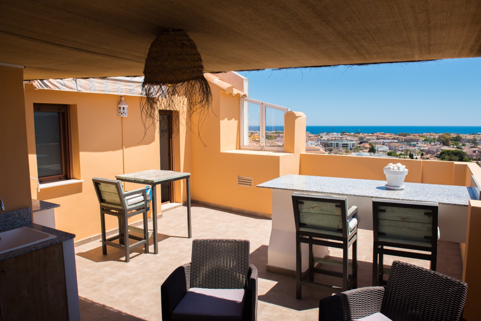 Terraza en la azotea con barra de granito, asientos de mimbre y vistas panorámicas al mar. Incluye zona de sombra, fregadero exterior y suelos cerámicos.