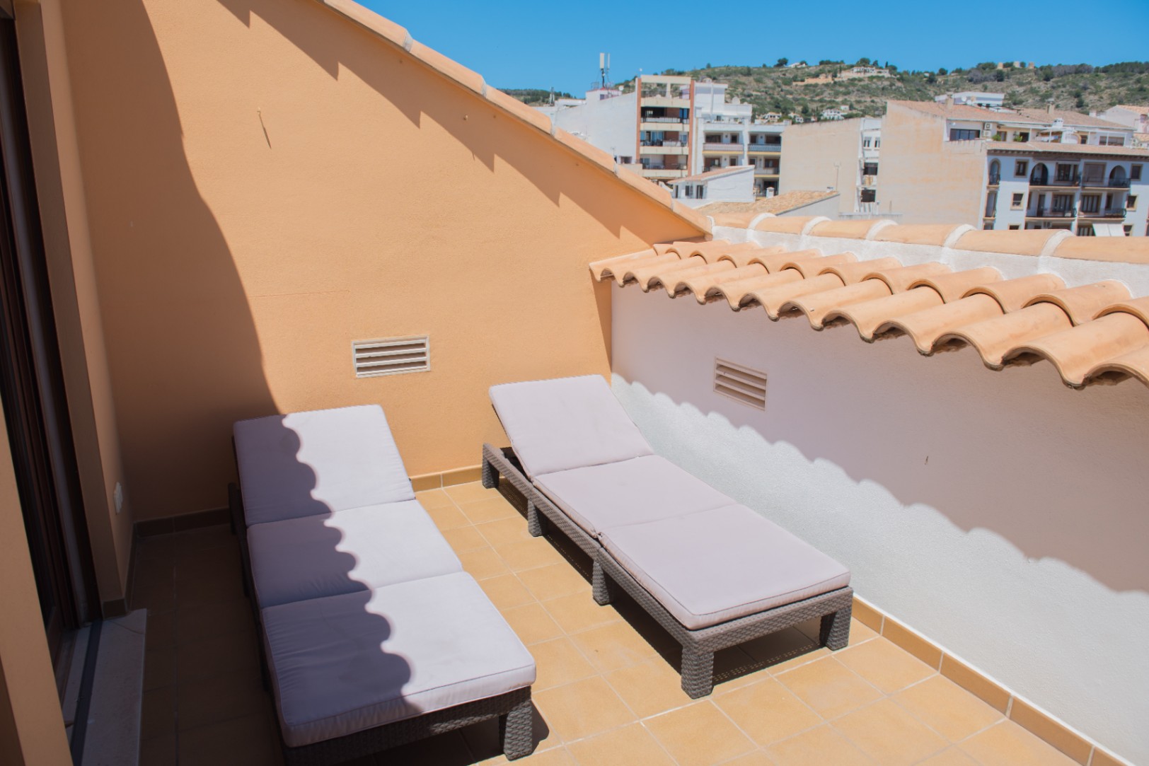 Terraza privada en la azotea con dos tumbonas de mimbre gris, suelo de baldosas de terracota y vistas al entorno urbano y las colinas.