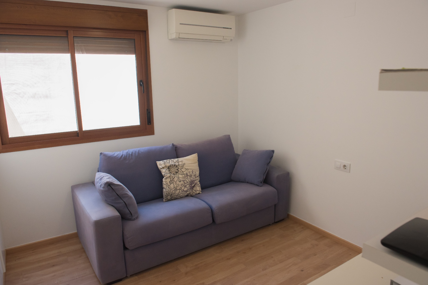 Zona de estar con sofá de tela azul, suelos de madera clara y aire acondicionado split junto a una ventana con marco de madera natural.