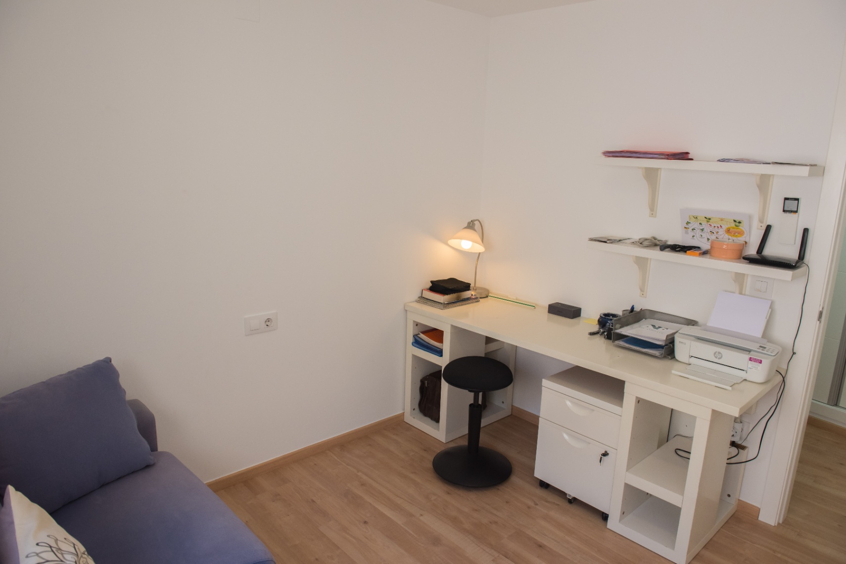 Zona de estudio funcional con escritorio blanco, almacenaje integrado y taburete negro ajustable sobre suelo de madera clara y paredes blancas.